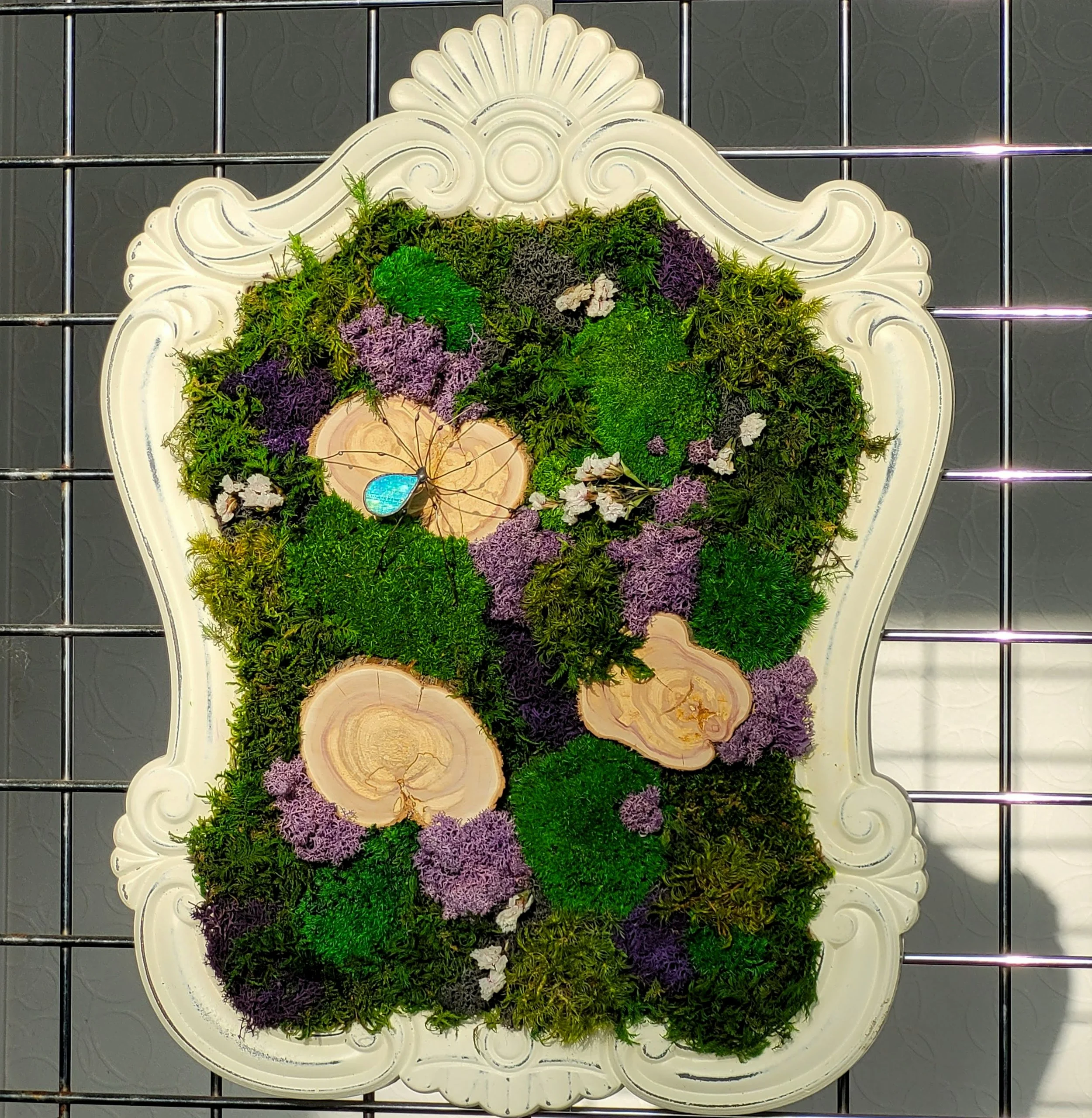Decorative wall art with green moss, purple flowers, wood slices, and a blue gem in an ornate ivory frame.