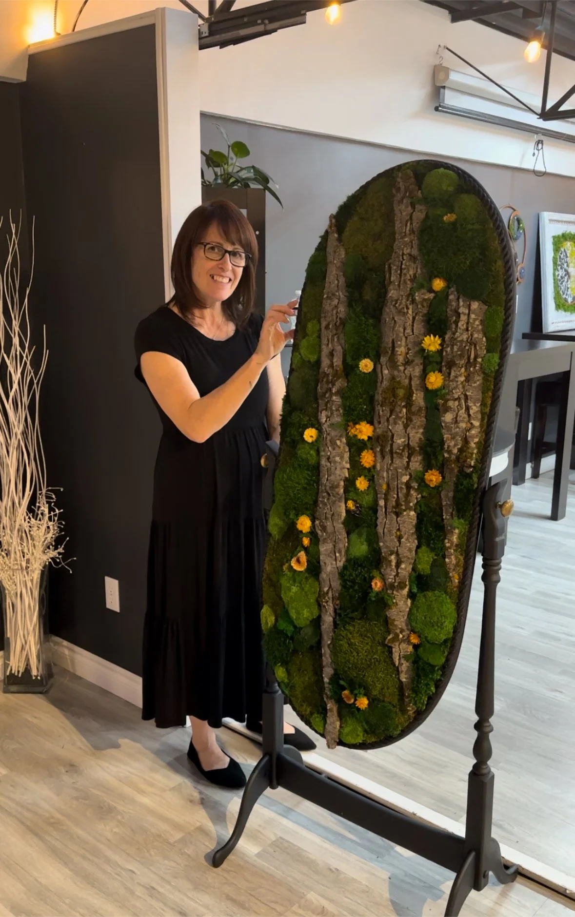 Woman standing next to a large, oval-shaped piece of wall art adorned with bark, moss, and yellow flowers in a modern room.