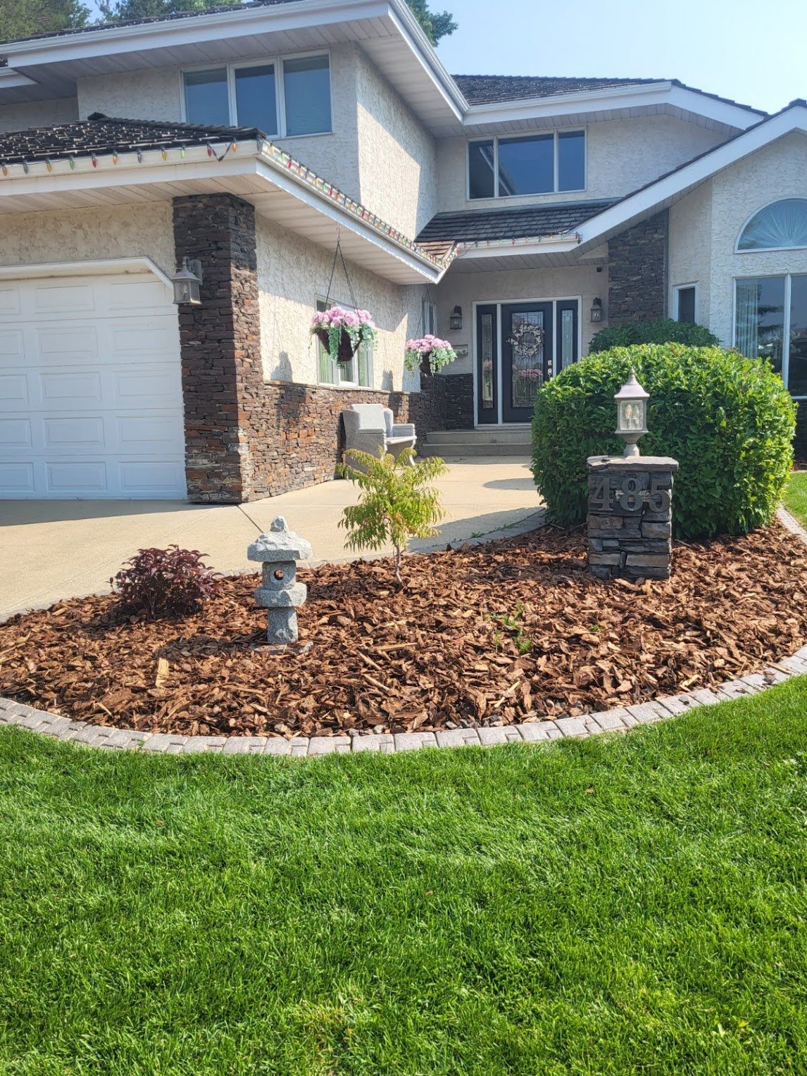 Suburban house front view with manicured lawn, stone landscaping, and a path leading to the entrance. The house features a gable roof, brick and stucco exterior, and decorative outdoor elements like a small garden lantern and pink hanging flowers nea