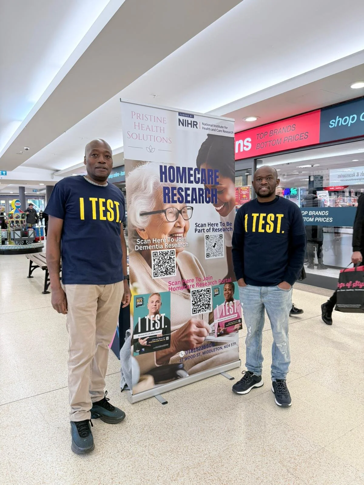 Two men wearing navy blue t-shirts with yellow text that reads "I TEST" stand on either side of a research poster in a shopping mall. The poster promotes home care research and features an elderly woman with white hair and glasses, and a young woman smiling. The poster includes QR codes and additional information about joining dementia research.