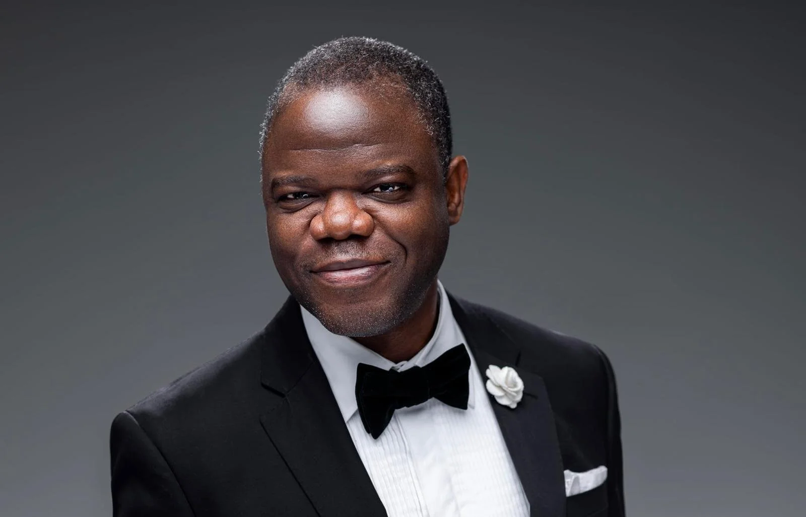 A middle-aged man in a tuxedo with a bowtie, white shirt, and a white flower on his lapel, smiling against a grey background.
