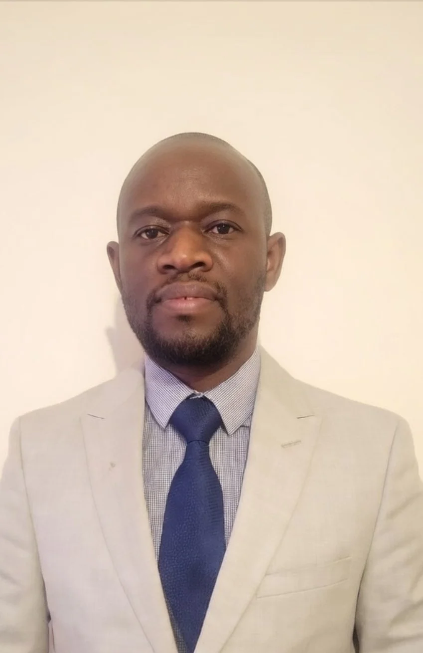 A man in a light-colored suit with a collared shirt and dark tie standing against a plain white wall.