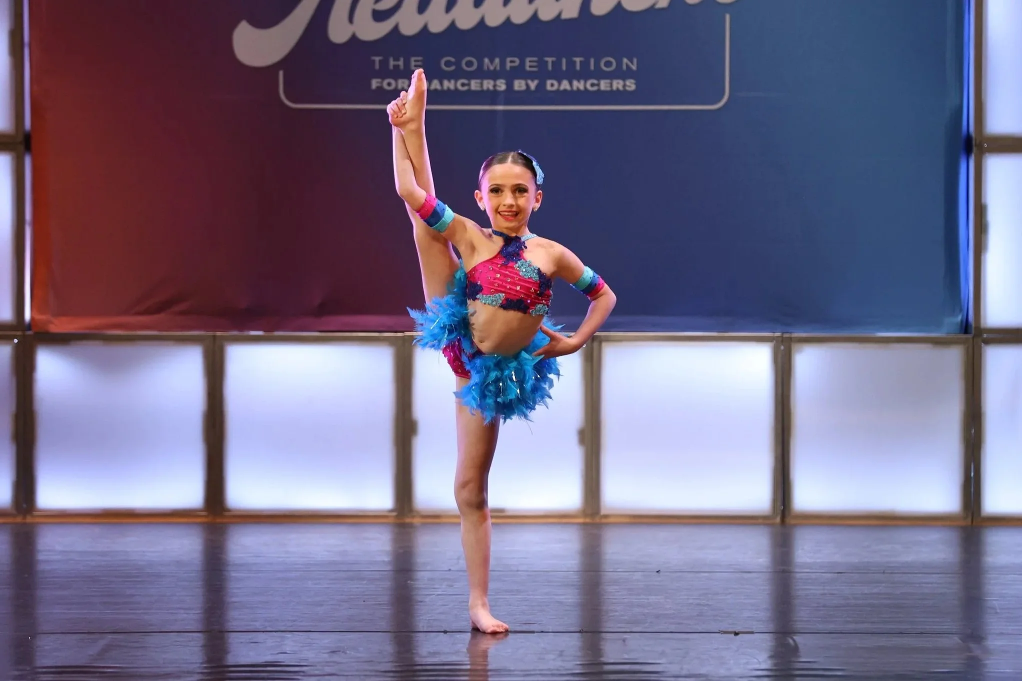 A young girl performing a ballet pose on stage during a competition, wearing a colorful costume with feathers, in front of a backdrop with the word 'Headliners' and smaller text.