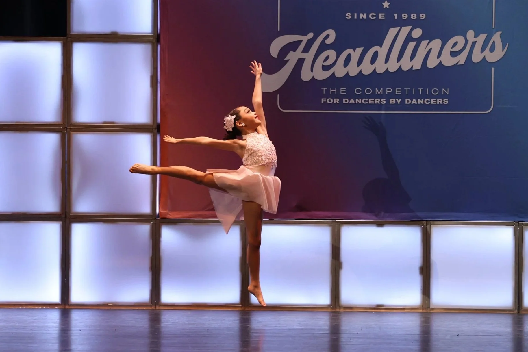 A ballerina performing on stage in front of a blue and purple backdrop with the words "Headliners" and "Since 1989."