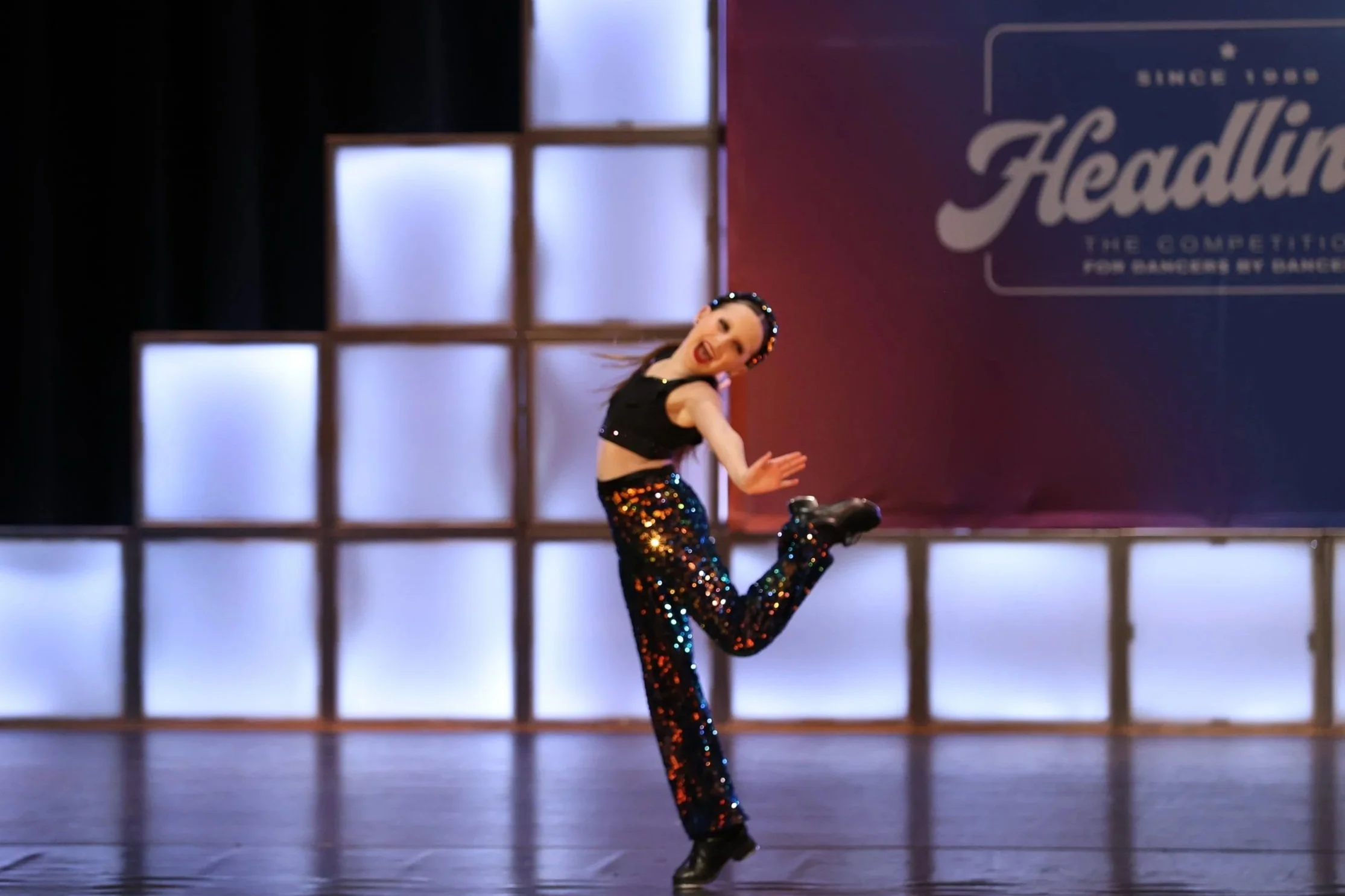A female dancer performing on stage wearing a black top, colorful sequin pants, and a black headband, with a backdrop that says 'Headline The Competition for Dancers by Dancers'.