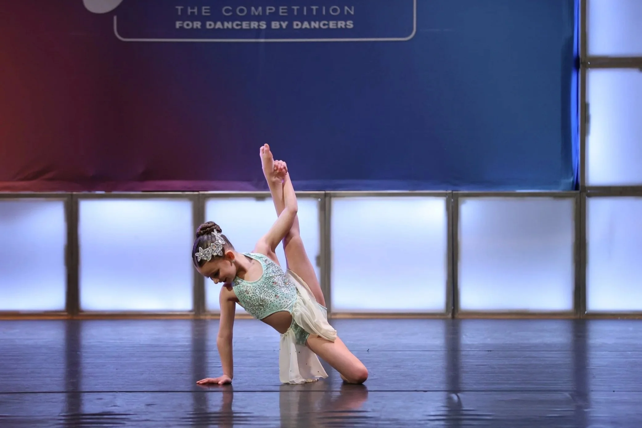 A young girl in a dance costume performing a pose on stage, with one hand and knee on the floor and one arm raised high.