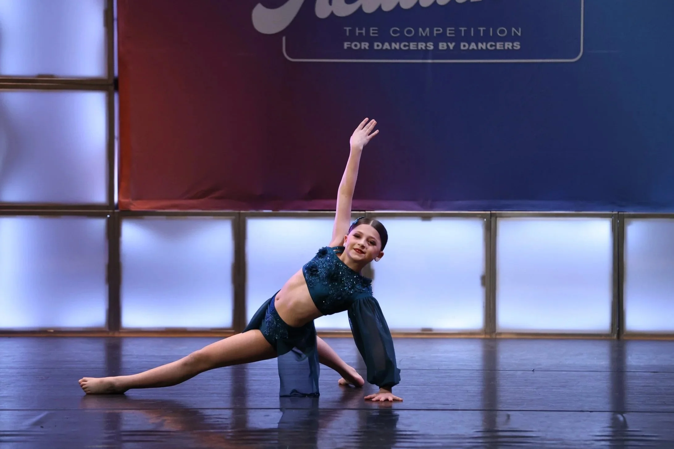 A young girl in a sparkly blue costume performs a dance move on stage during a dance competition, with a large banner in the background reading 'Hematite The Competition For Dancers By Dancers.'
