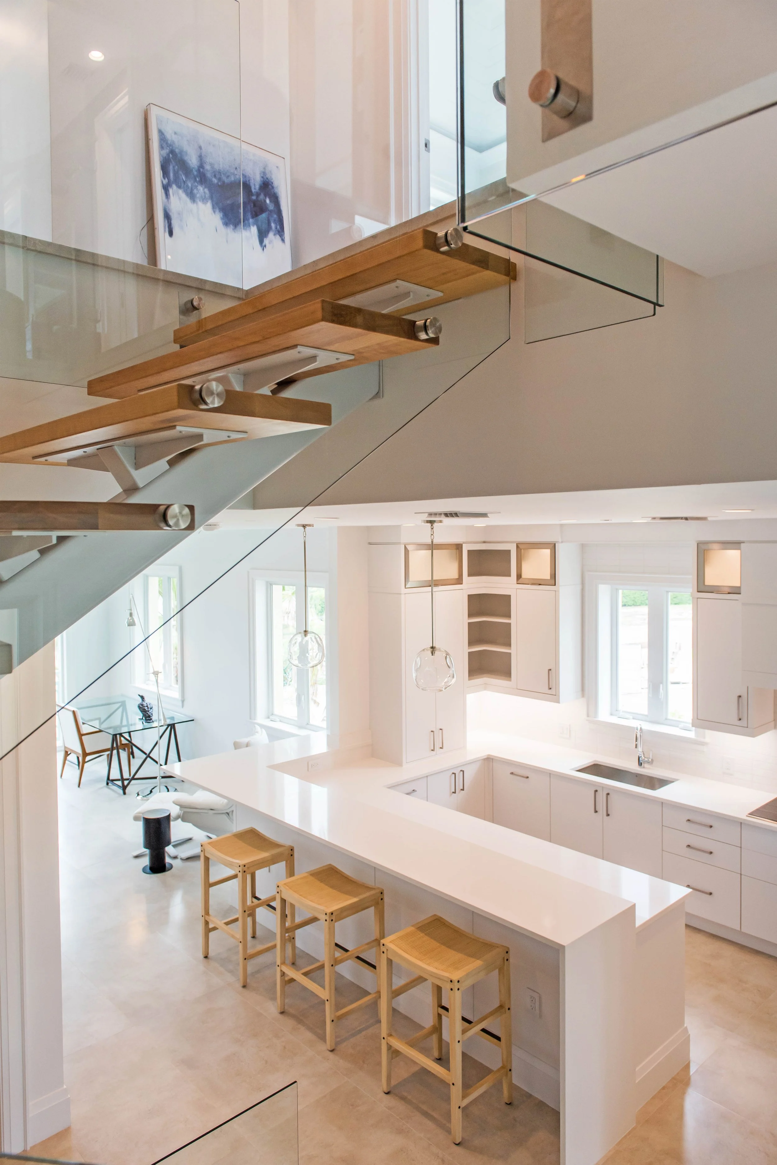 Modern kitchen with white cabinets, a large white island with three wooden stools, and large windows letting in natural light. There are two pendant lights above the island and a staircase with glass railing overhead.