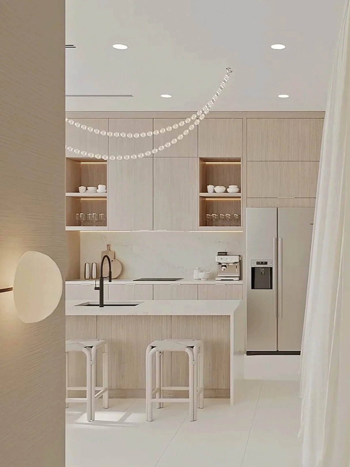 Modern minimalist kitchen with beige wood cabinets, white marble countertops, a black faucet, built-in refrigerator, and two white bar stools at the kitchen island. Pendant lighting and decorative shelves with white dishes and glasses