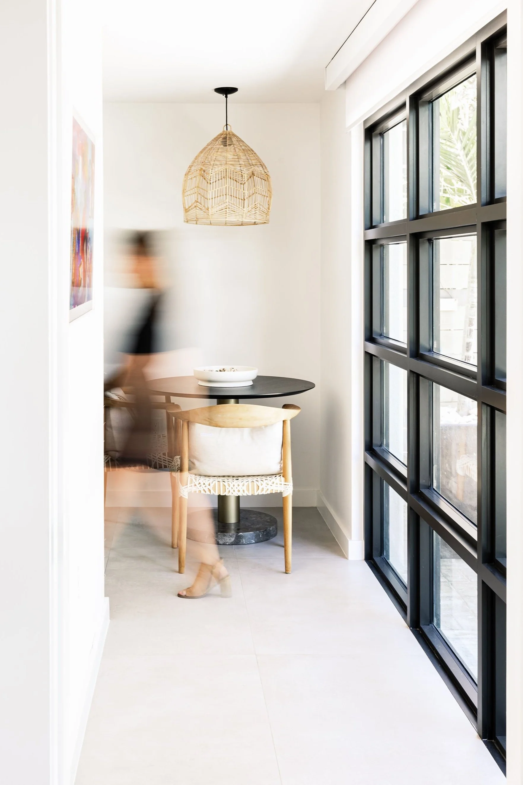 A blonde woman in black clothing walking past a small dining nook with a black round table and two chairs, one wooden with a woven seat, in a bright room with large grid windows and a rattan pendant light.
