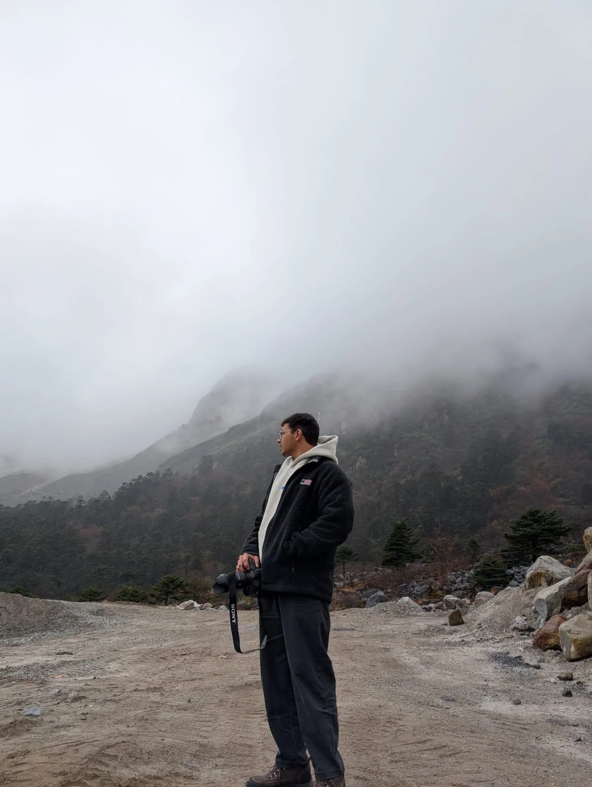 A man stands on a dirt path holding a camera, overlooking a foggy, mountainous landscape with trees and rocks.