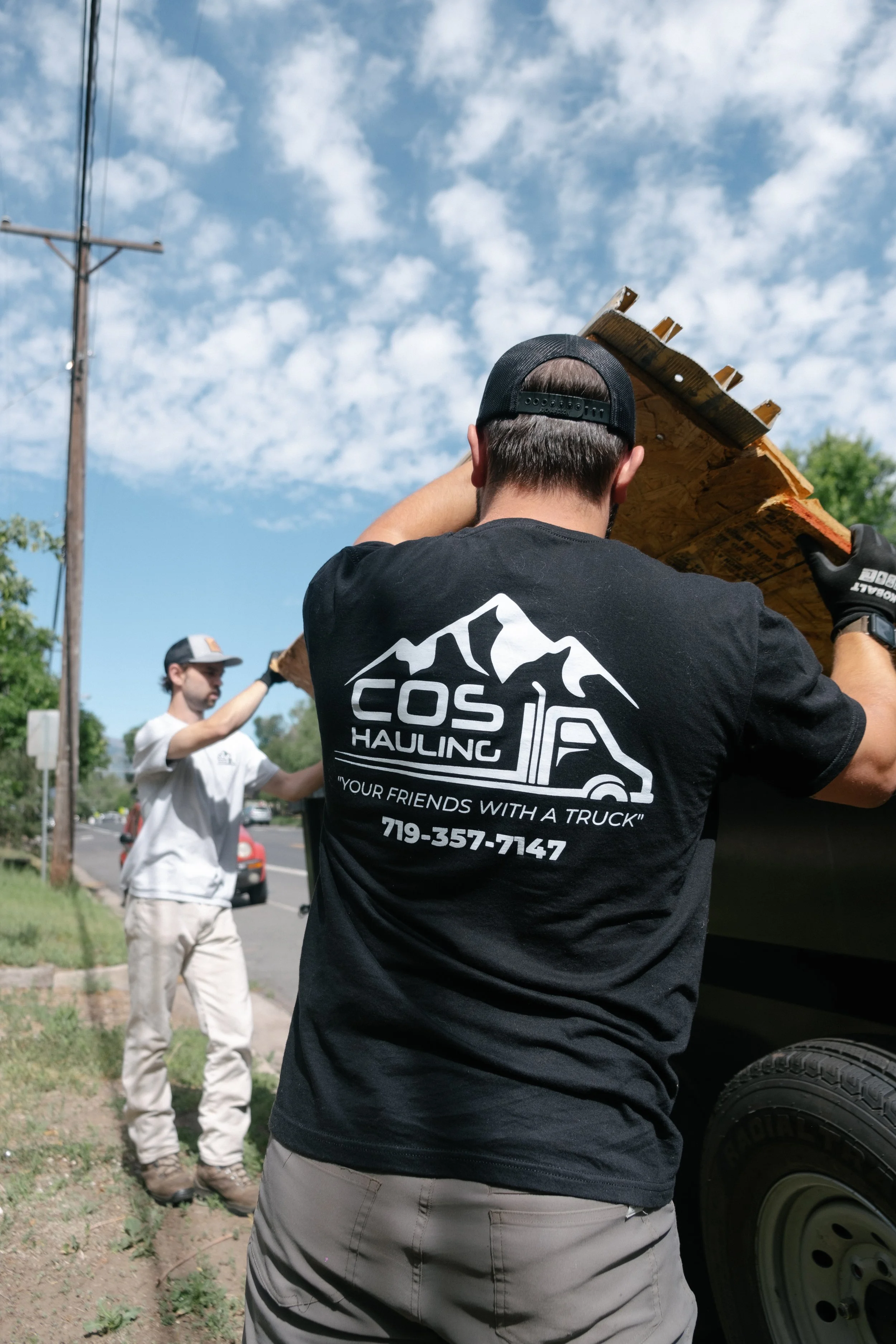 Two men are moving a large piece of wood or plywood. One man, wearing a black shirt and cap, is seen from behind, while the other man, in a white shirt and cap, is in the background. They are outdoors on a sunny day with a blue sky and clouds.