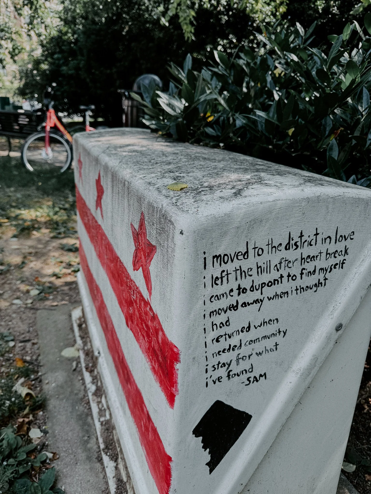 Close-up of a painted concrete block with handwritten text, a red arrow, and black paint marks, set outdoors with greenery and a bicycle in the background.