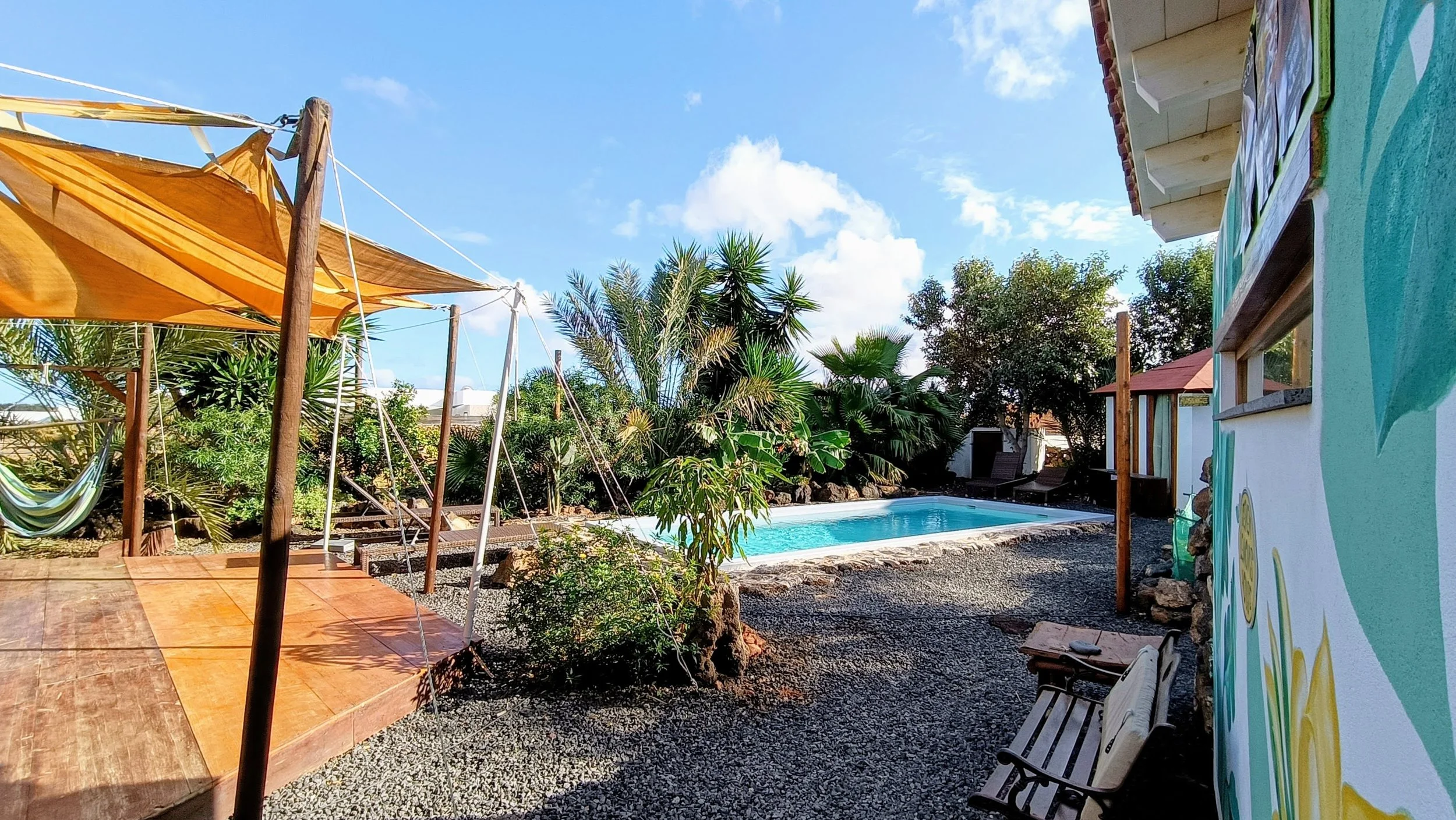 A backyard with a swimming pool, trees, and seating area. There is a wooden deck with a canopy on the left, garden chairs on the right, and a colorful wall mural on the right side of the house. The sky is blue with some clouds.