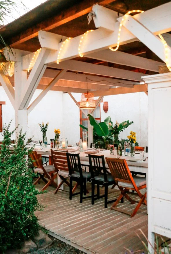 Outdoor dining area with a long table set for a meal, surrounded by wooden chairs. The table has flowers, bottles of water, glasses, and candles. There are plants and greenery in the background, and string lights and a hanging light fixture decorate 