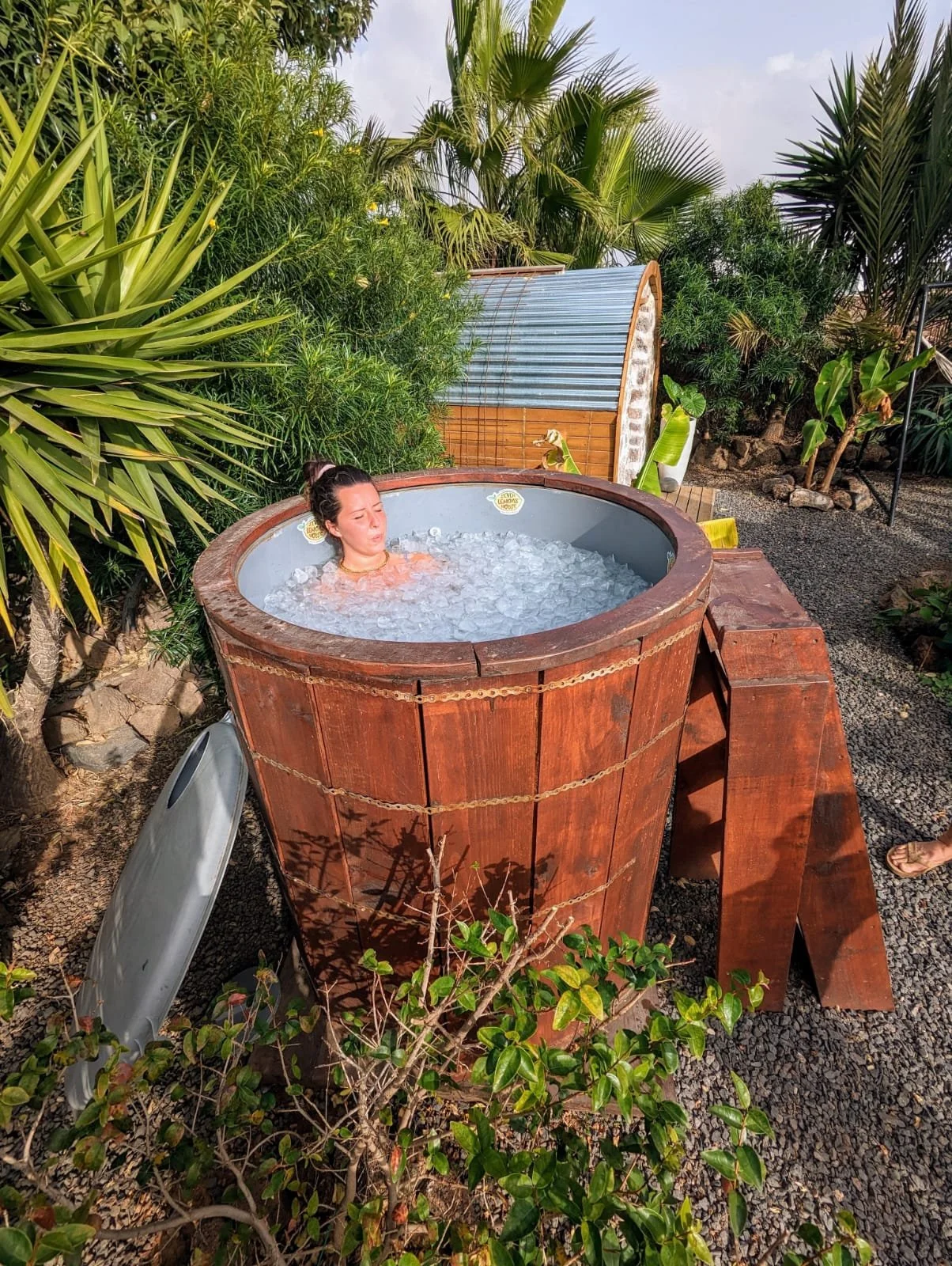 Woman relaxing in a wooden hot tub outdoors surrounded by greenery and tropical plants.