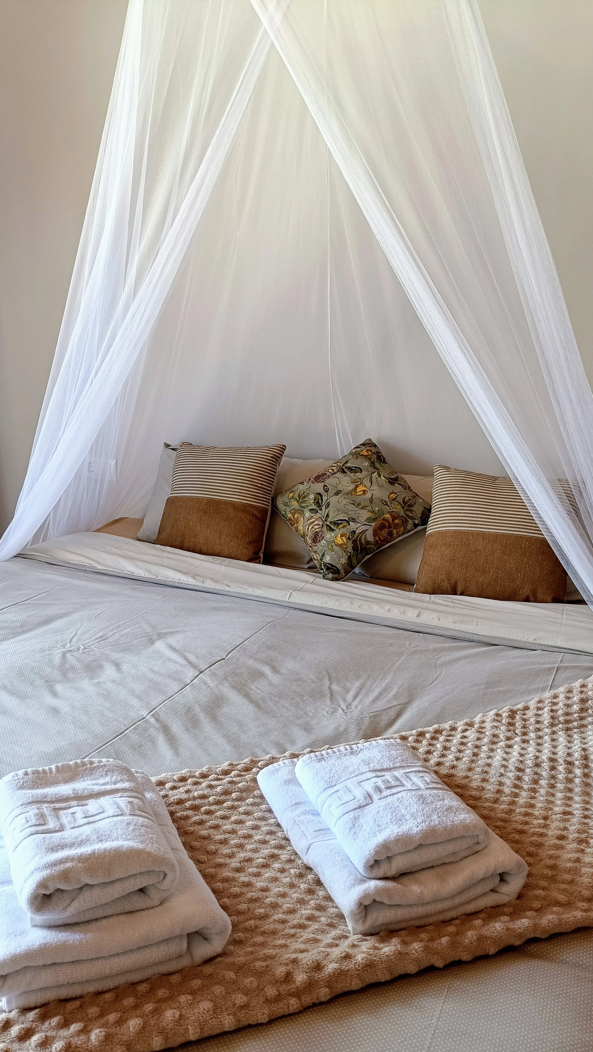The image shows a neatly made bed with white linens, topped with four decorative pillows, including one with a floral pattern. White sheer curtains hang above the bed, creating a canopy effect. In the foreground, there are four rolled white towels pl