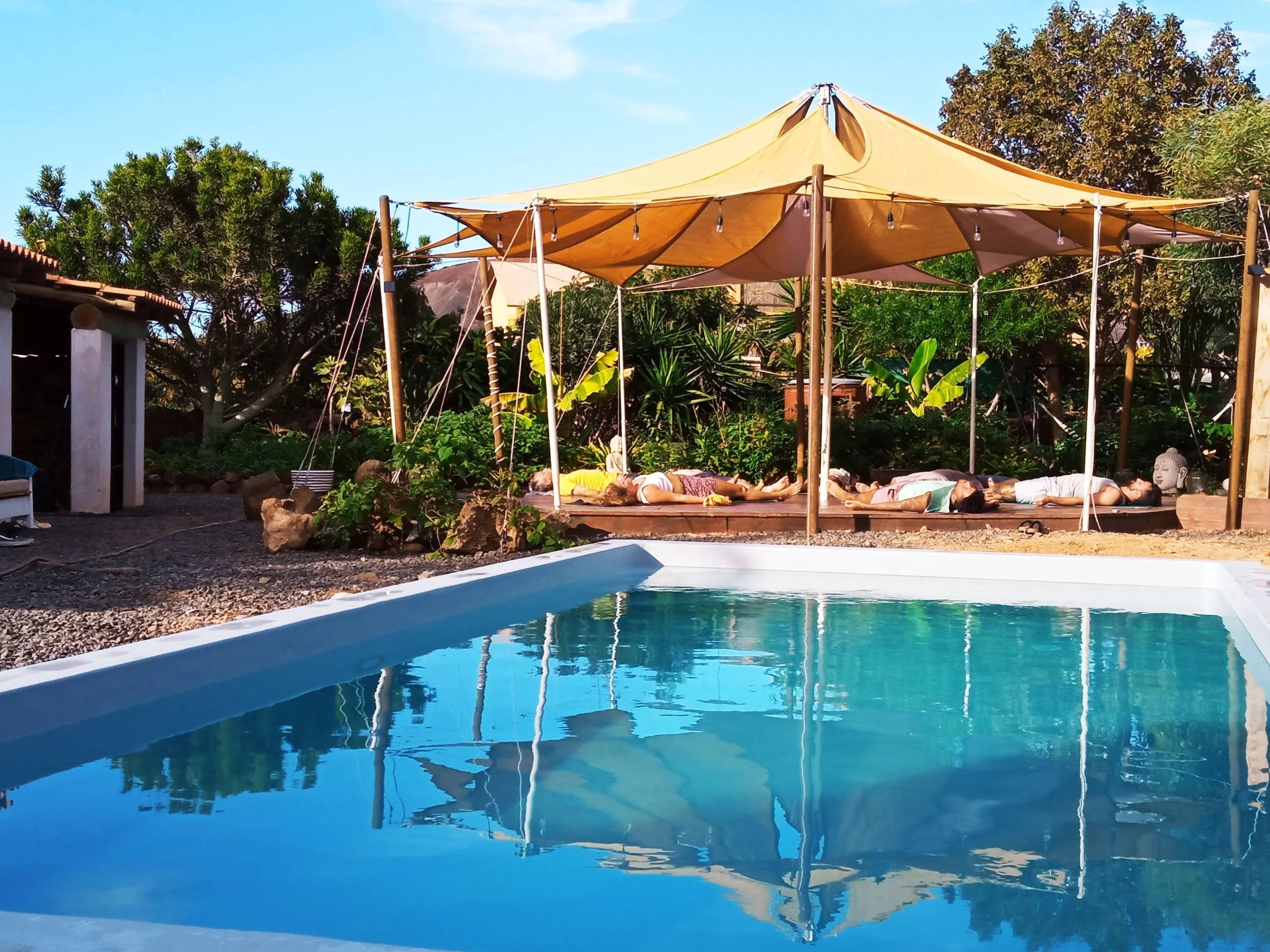 People practicing yoga on a wooden platform under a yellow canopy near a swimming pool, with trees and blue sky in the background.