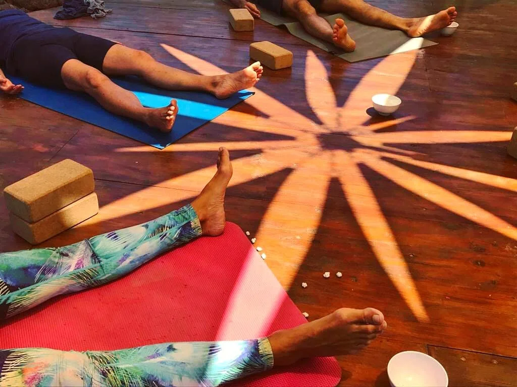 People practicing yoga in a studio with wooden floors, using mats, blocks, and bowls, with sunlight casting a star-shaped shadow on the floor.
