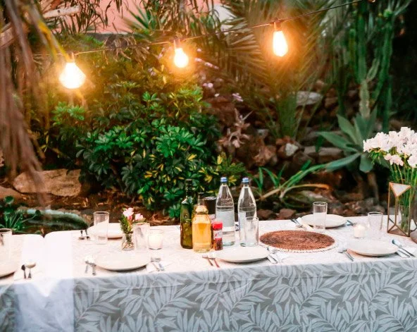 Outdoor dining table set for a meal with candles, bottled water, and drinks, surrounded by lush greenery and warm hanging lights.