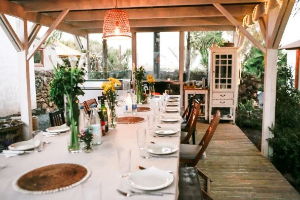 Outdoor dining table set with plates, glasses, bottles, and flowers under a covered patio with wooden flooring and greenery.