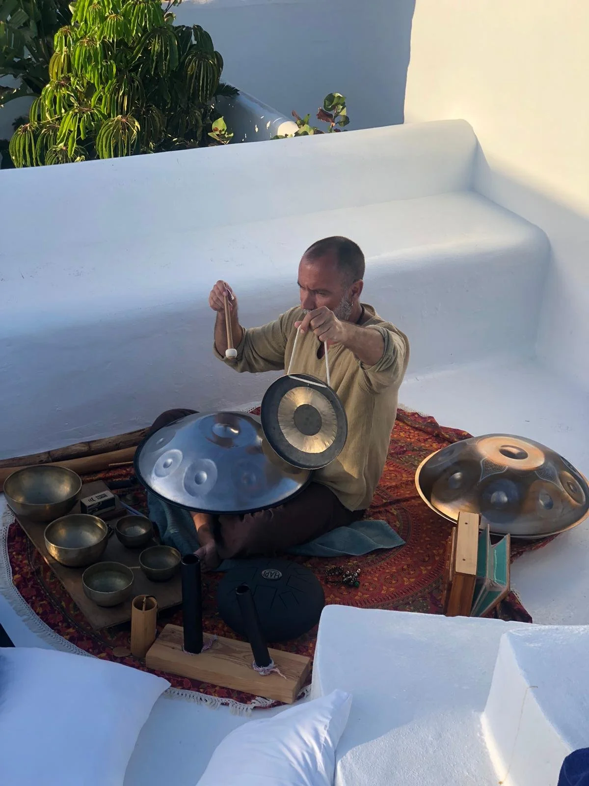 A man playing musical instruments, including a hang drum and a gong, on a decorative rug outdoors with white walls and green plants in the background.