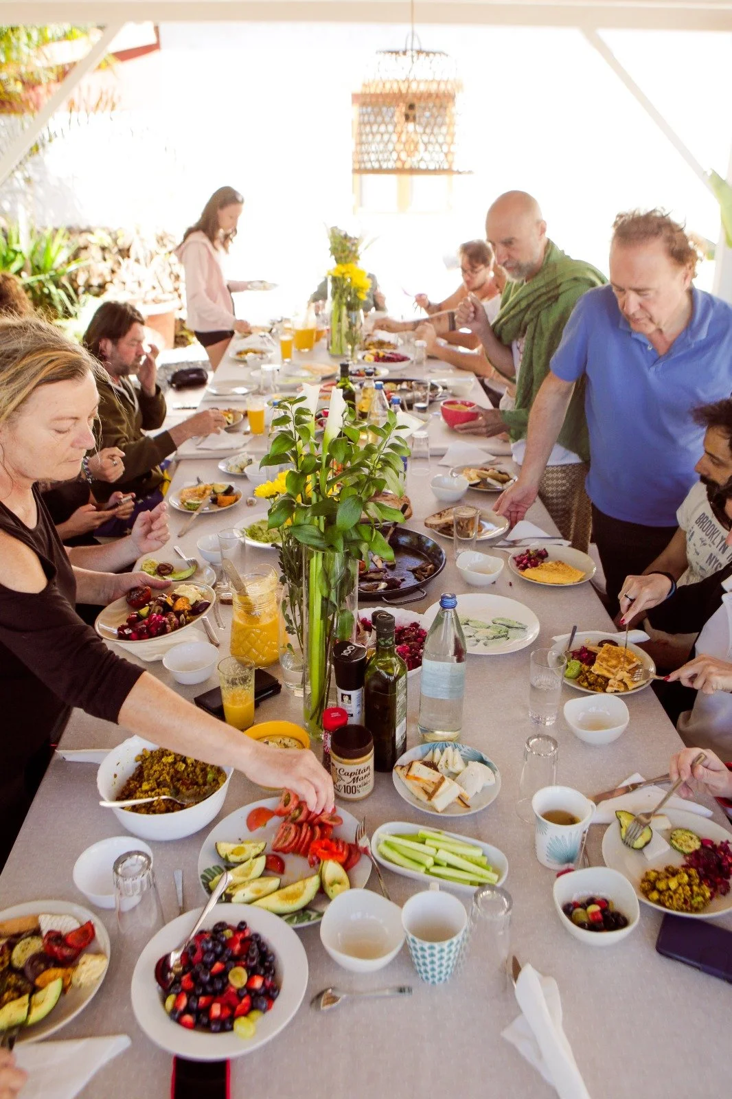 A large outdoor gathering with people sitting and standing at a long table enjoying a meal, with plates of food, drinks, and a central plant decoration.