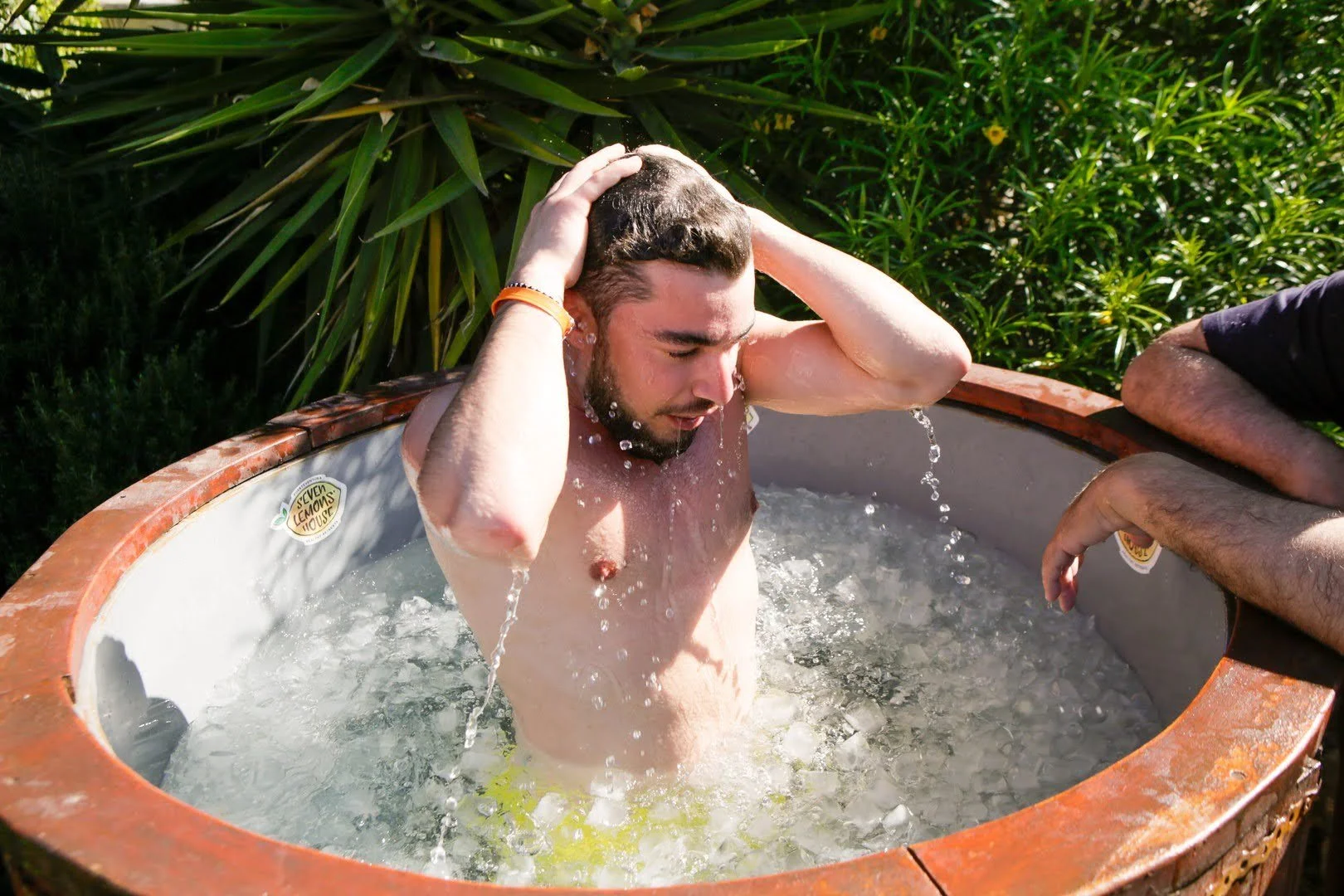 A shirtless man with dark hair and a beard washing his hair in an outdoor tub surrounded by green plants.