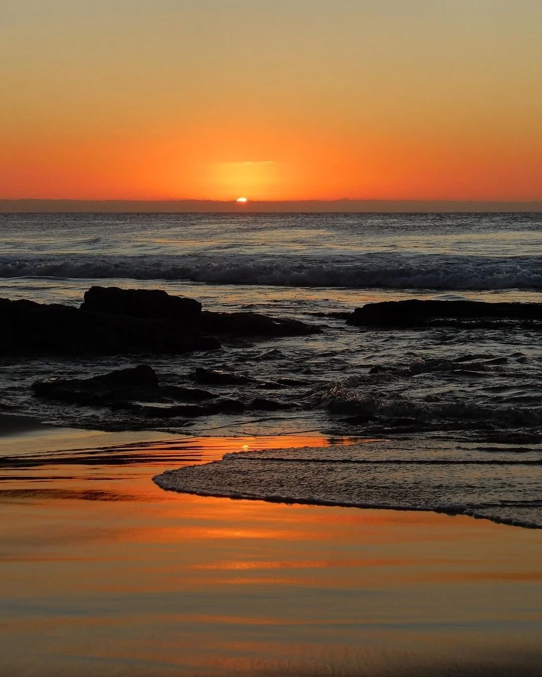 A serene beach at sunset with orange and yellow sky, gentle waves, rocks in the foreground, and a reflection of the sunset on the wet sand.