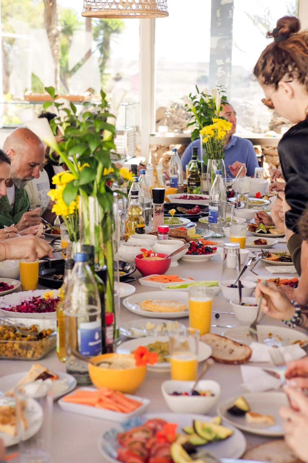 People gathered around a long dining table filled with breakfast foods, flowers, and drinks, in a bright room with large windows and greenery outside.