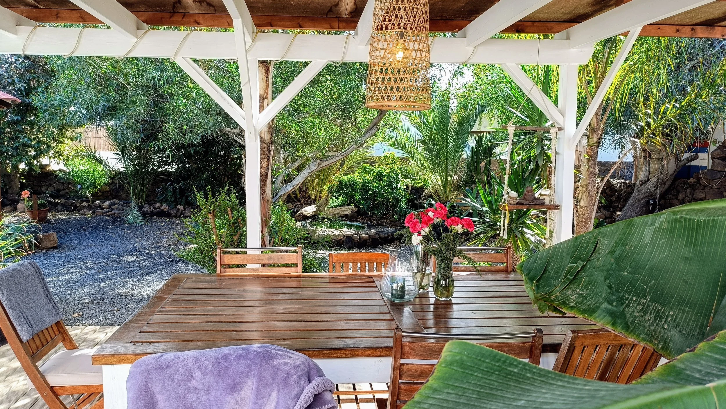 Outdoor patio with a wooden dining table, surrounded by chairs, under a wooden roof with white support beams. There are vases with pink and white flowers on the table. The background features lush greenery with trees and plants, creating a garden sce