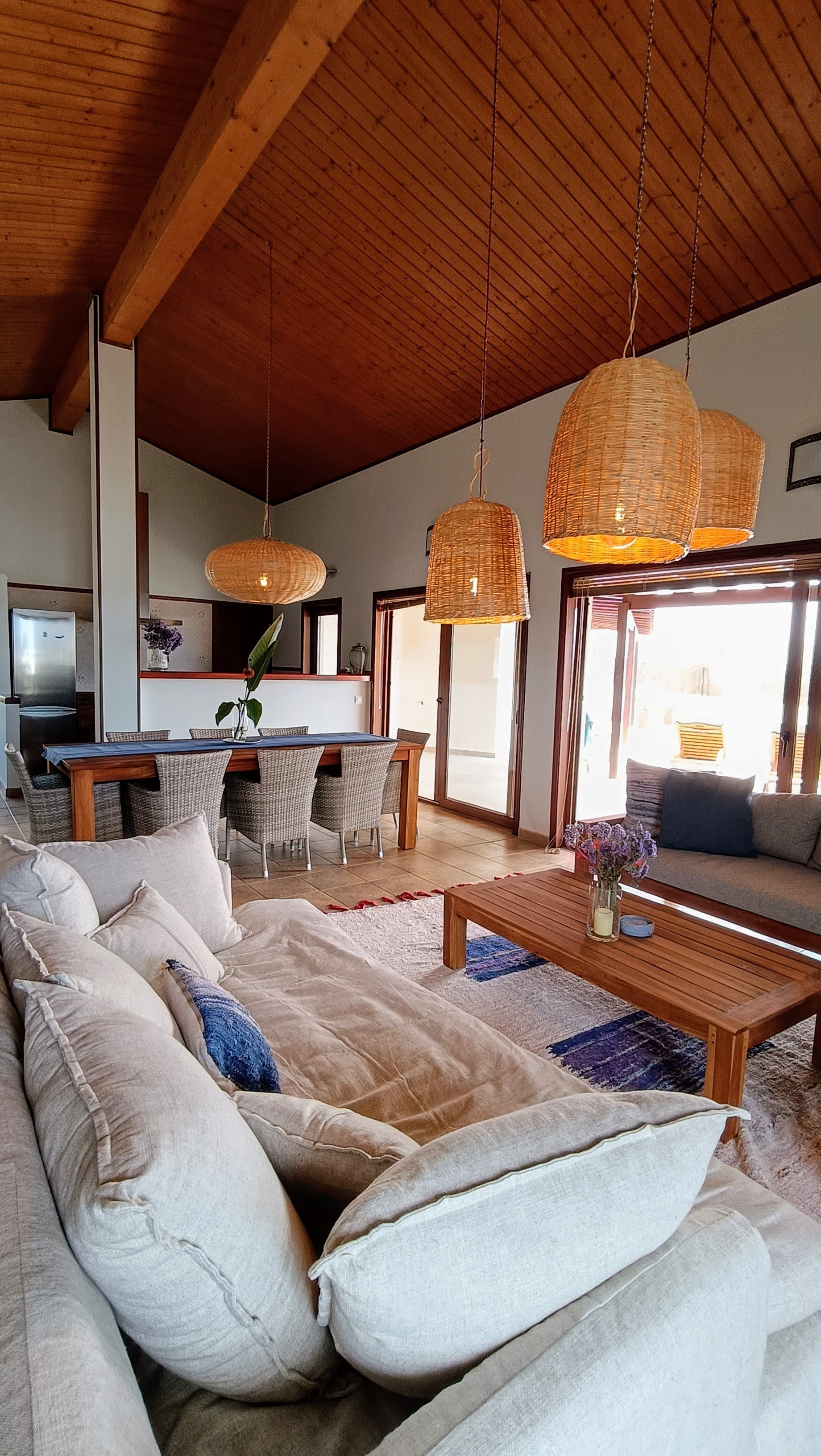 Living room with beige sofa, wooden coffee table with purple flowers, dining table with wicker chairs, pendant lights hanging from wooden ceiling, sliding glass door leading outside.