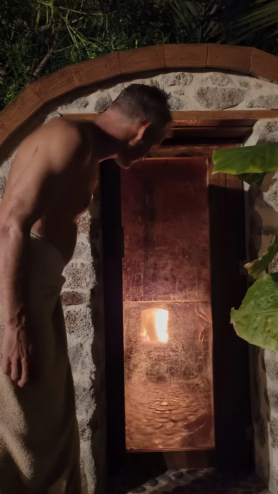 A shirtless man looking into a fire-lit enclosed space, surrounded by greenery and stone wall