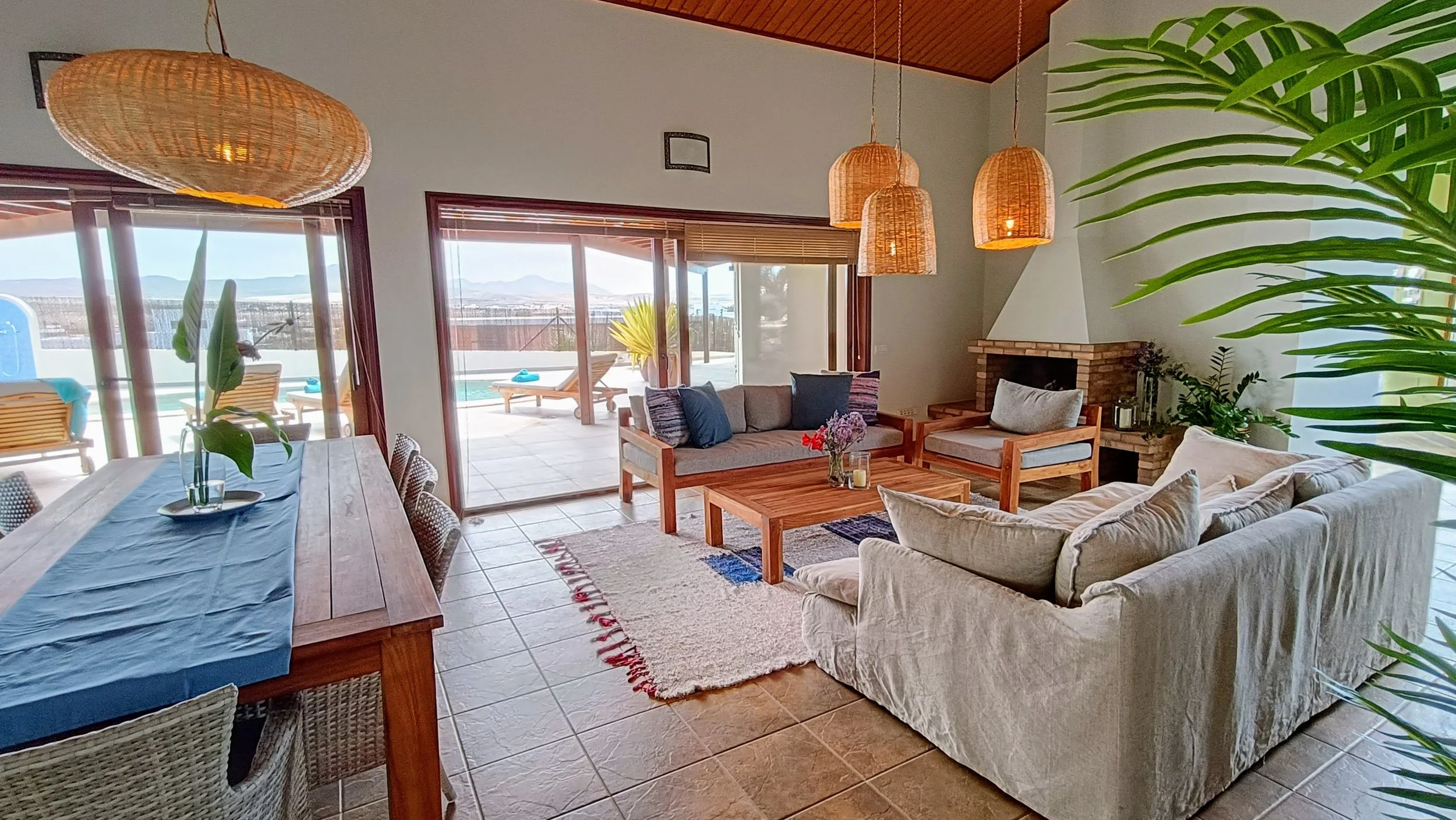 Living room with beige sofa, wooden coffee table with flowers, and a fireplace, with sliding glass door leading to patio with lounge chairs and a hot tub, overlooking landscape and mountains.