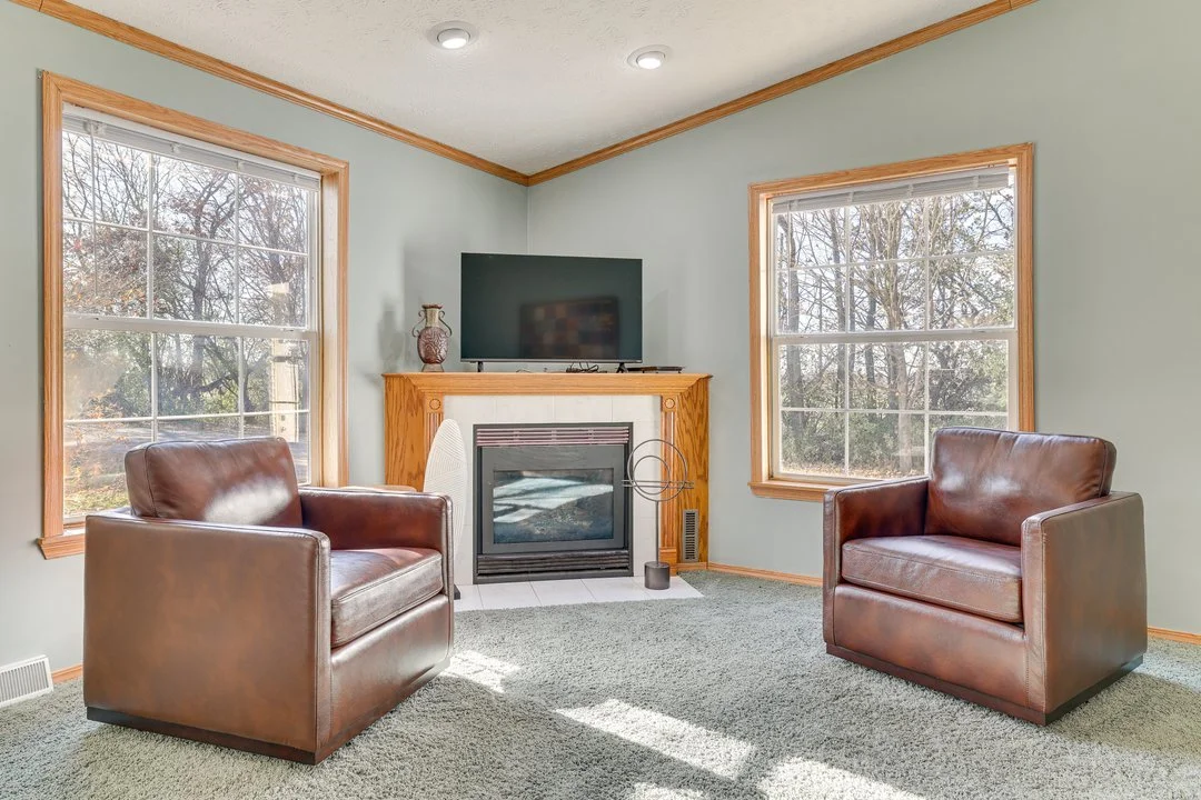 Living room with two brown leather chairs, a fireplace with a wooden mantel, a TV on top, large windows showing trees outside, and carpeted floor.