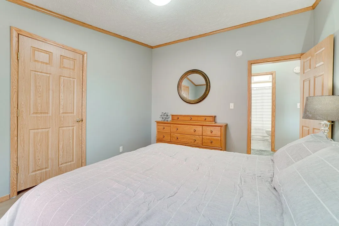 A bedroom with light green walls, a wooden dresser with a mirror above it, a bed with white bedding, and an open door leading to a bathroom.