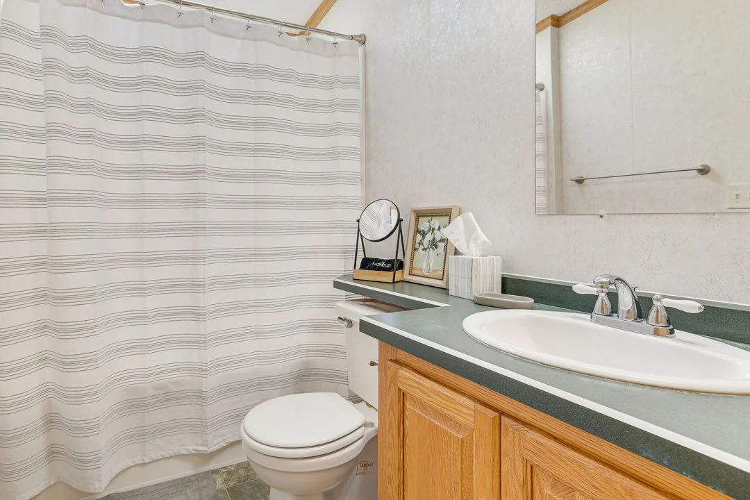 Bathroom with a shower curtain, toilet, wooden vanity with a white sink, mirror, tissue box, framed picture, and decorative items.