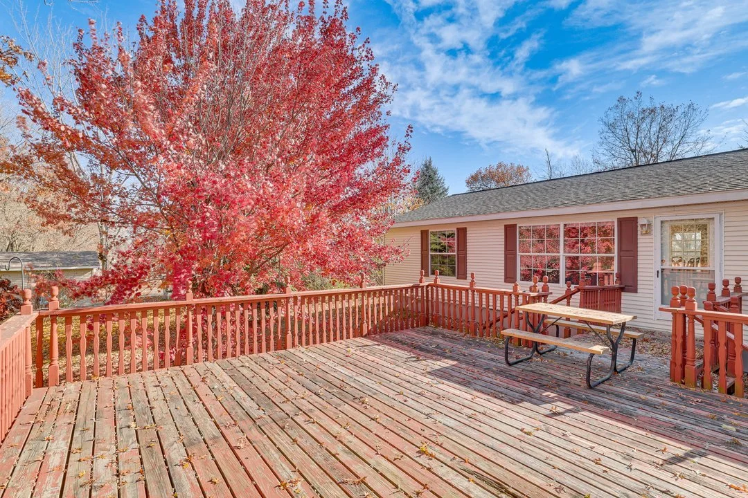 A wooden deck attached to the house with brown shutters and a glass door, overlooking a large red and pink flowering tree under a blue sky with some clouds.