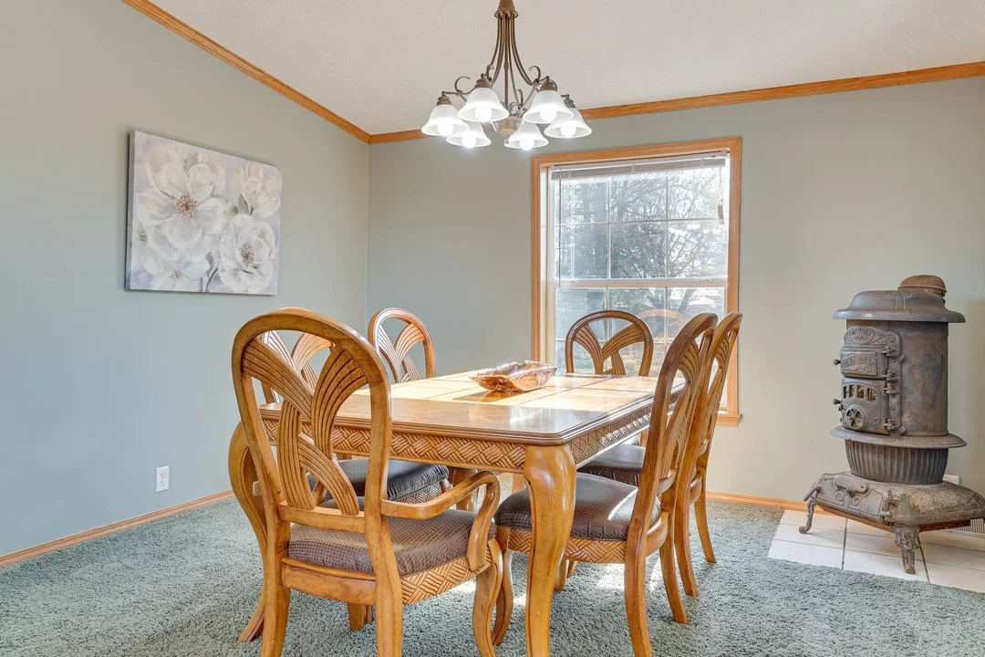 Dining room with a wooden table and six chairs, a large window, a chandelier, a wall painting of white flowers, and an antique wood stove in the corner.