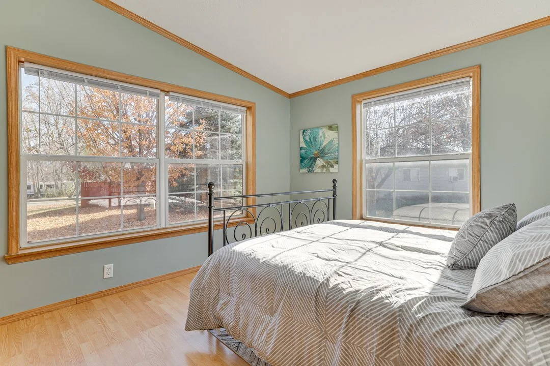 A bedroom with a metal bed frame, light green walls, light wooden window frames, and two large windows revealing a yard with trees outside. The bed has striped pillows and a duvet.