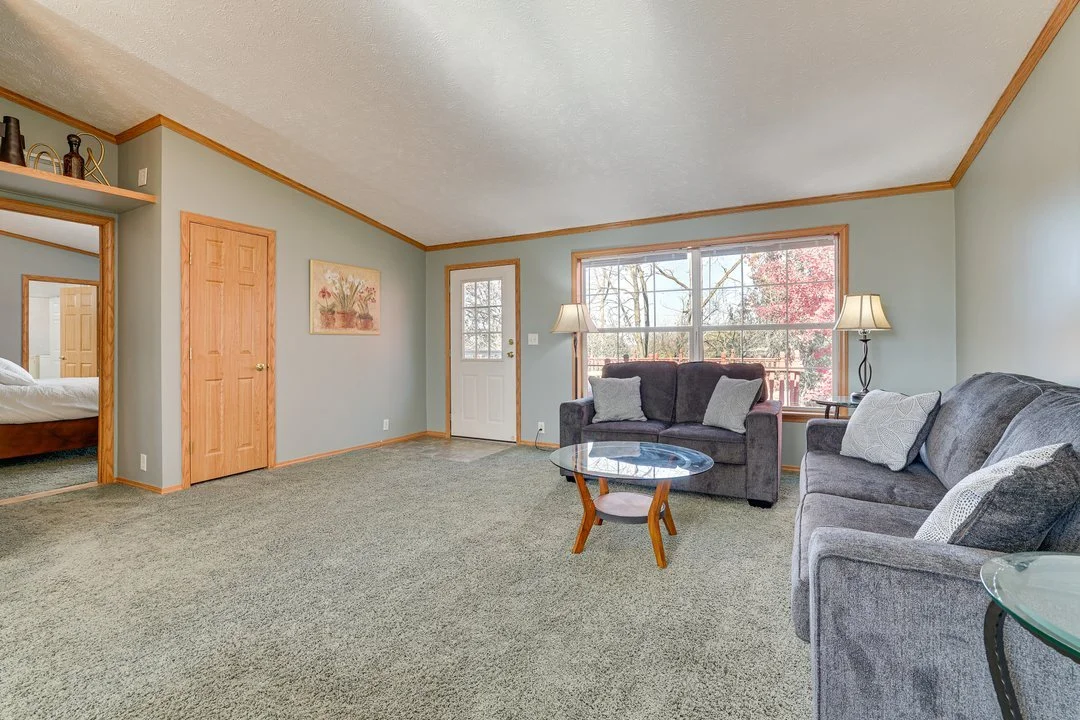 Living room with light green walls, large window, gray sofas, glass coffee table, and lamps, with door leading outside and door to bedroom