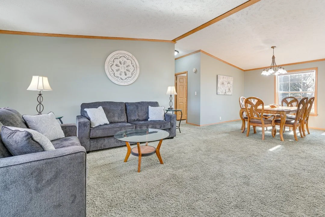Living room with light green walls, gray sofas with patterned pillows, a wooden coffee table with glass top, side tables with lamps, and a dining area with a wooden table and chairs near a large window.
