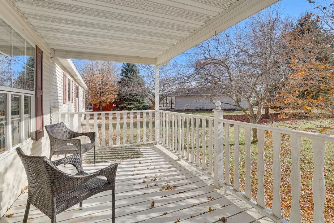 A sunny porch with two wicker chairs and a white railing, overlooking a yard with trees and fallen leaves in autumn.