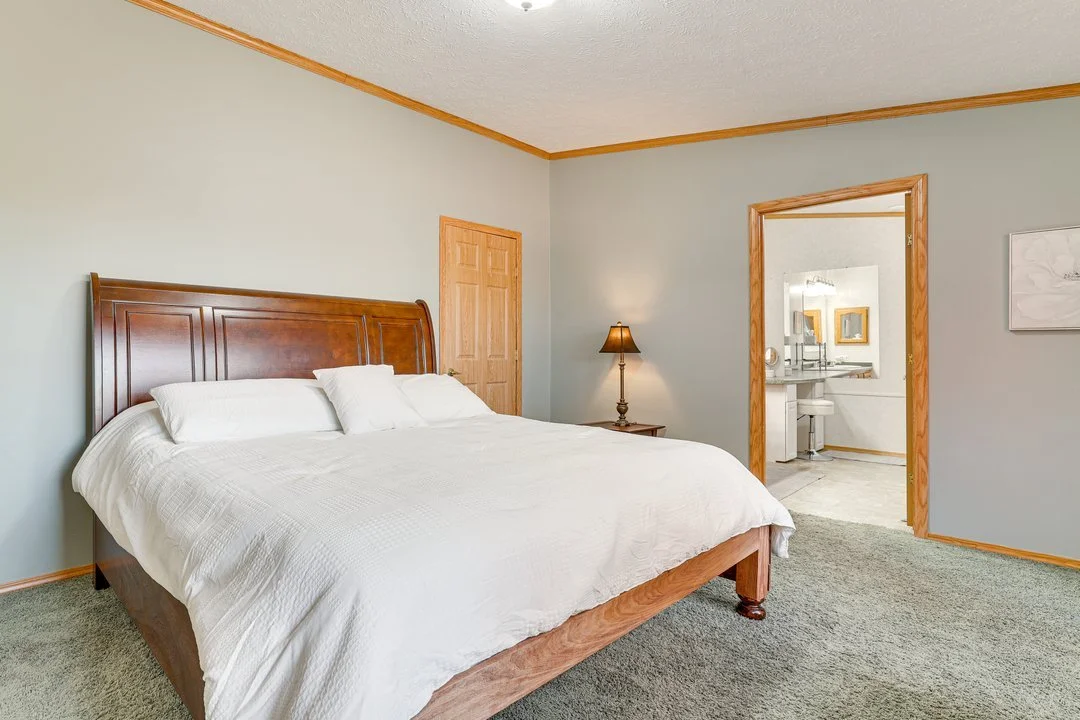A bedroom with a large bed with white bedding, a wooden headboard, a nightstand with a lamp, and a doorway leading to a bathroom.