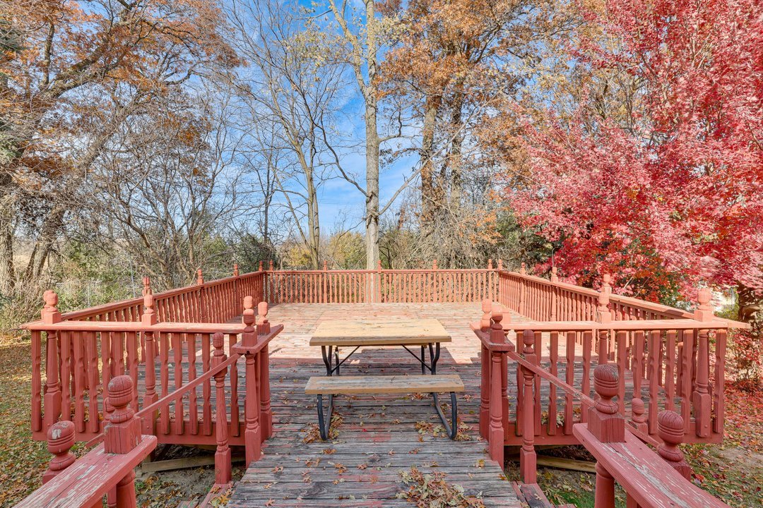 An outdoor wooden deck with a picnic table, surrounded by red, orange, and brown autumn trees under a partly cloudy sky.