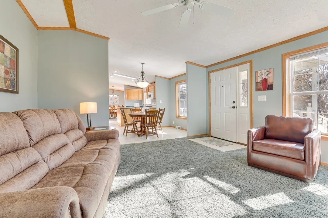 Living room with beige sofa, brown leather armchair, green carpet, patterned wall art, and large windows; view into kitchen and dining area with wooden furniture and chandelier.