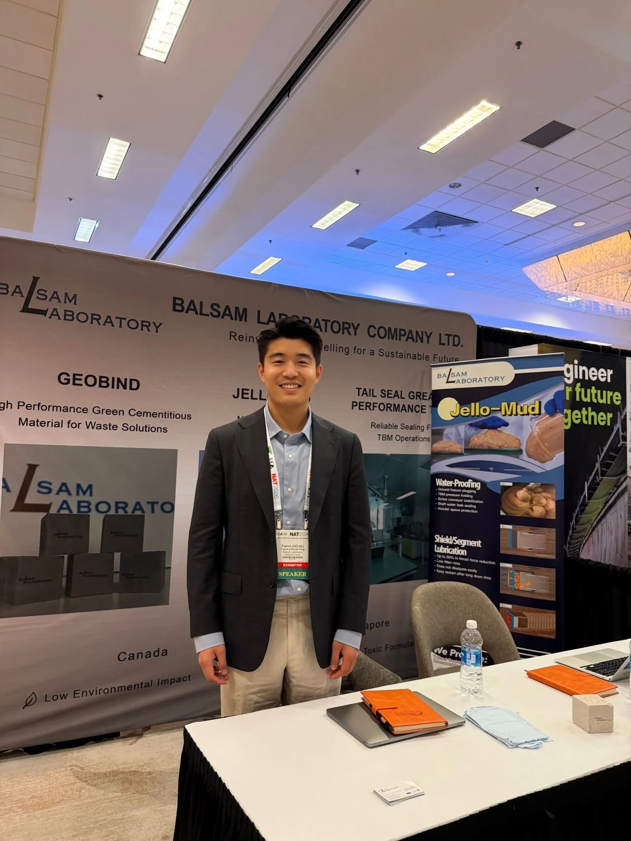 A man in a suit standing at a conference booth with banners displaying information about Balsam Laboratory products like Jello-Mud and Sealant. There are tables, notebooks, a water bottle, and chairs in the foreground.