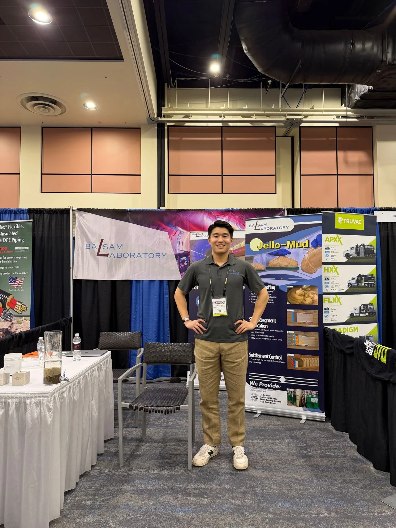 A young man standing and smiling at a trade show booth for BLSAM Laboratory, with posters and banners displaying information about construction materials and services.