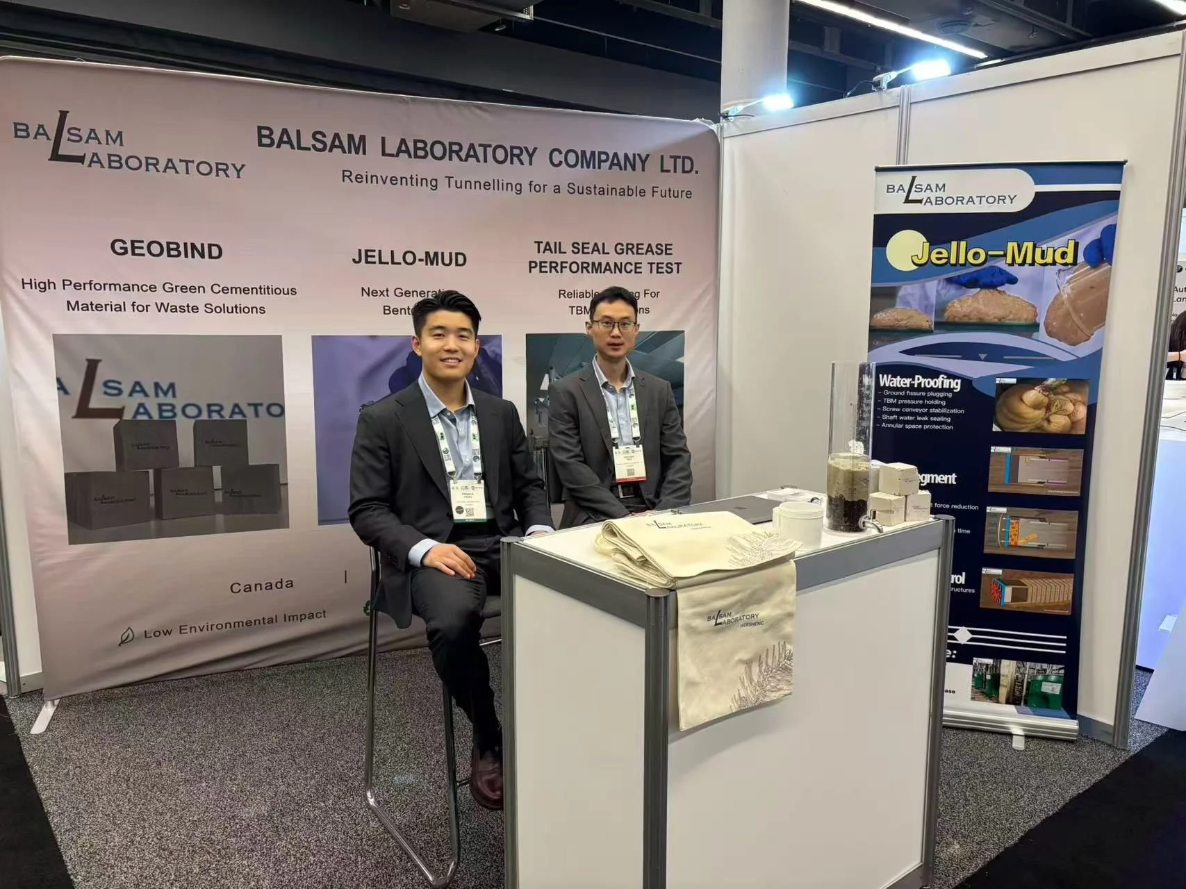 Two men in business suits sitting at a booth display at a trade show. The booth is for BALSAM Laboratory, showcasing their products and research related to tunneling technology and waterproofing materials. The background features banners with text about the company's innovative solutions for tunneling and waste management.