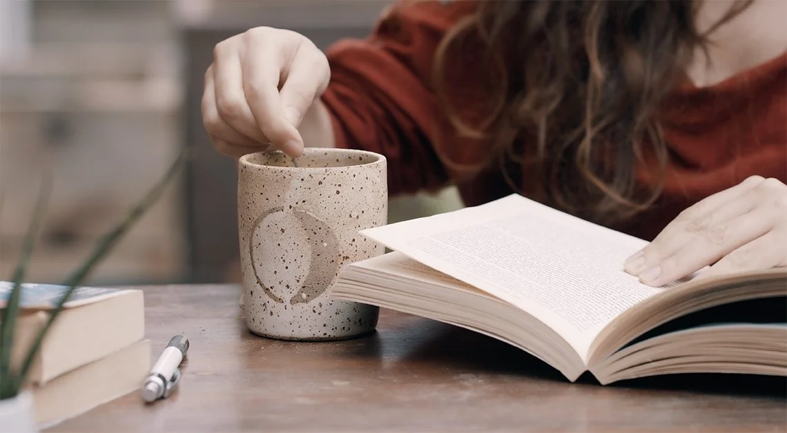 Image of a person reading a book at a desk whilst stirring a cup of coffee
